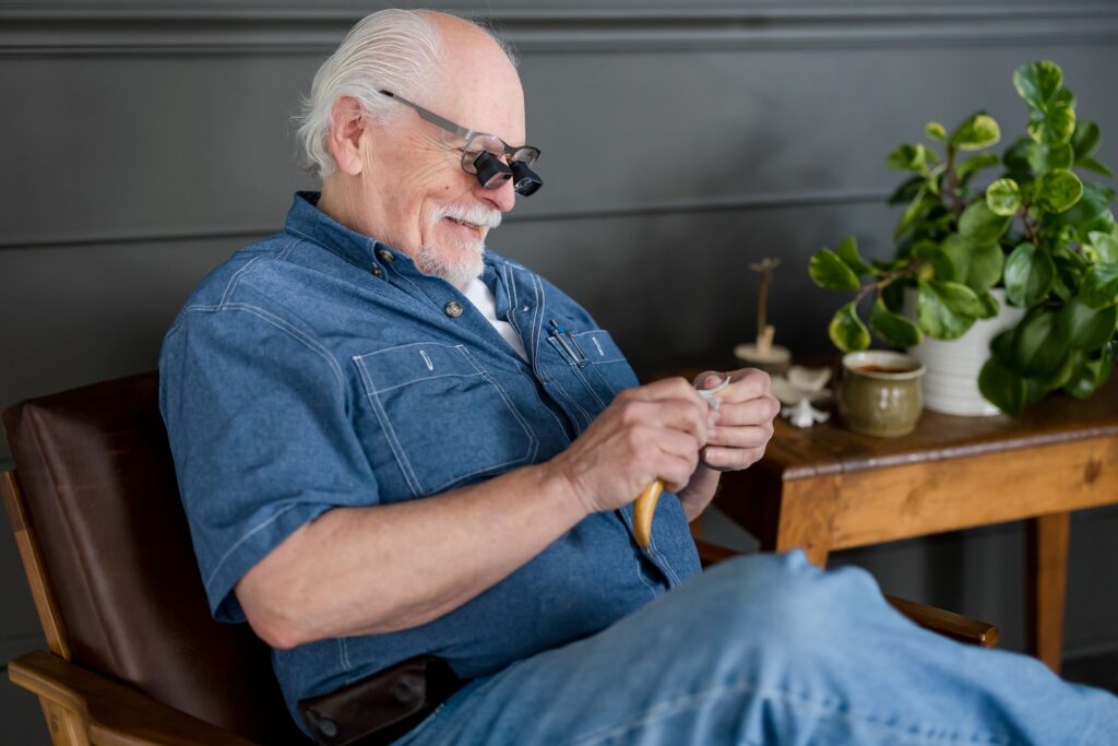 Man wearing low vision glasses sitting in a chair whittling wood