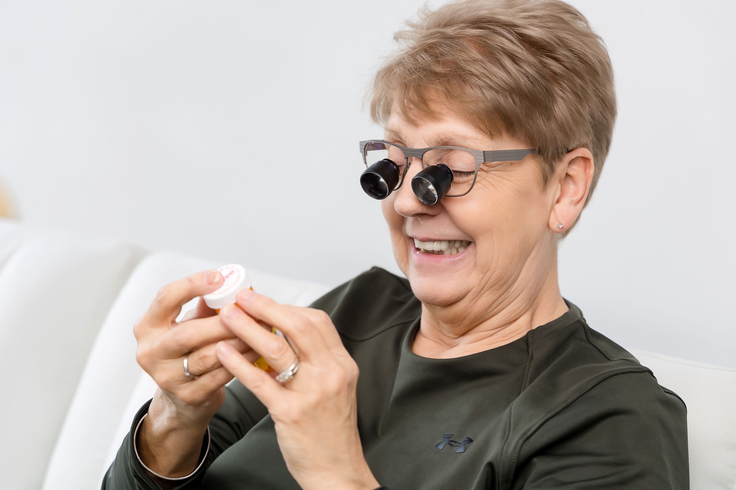 A woman wearing low vision glasses smiling looking at a prescription bottle