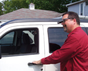 A man reaching for a door handle of a white car. He is wearing bioptic telescope glasses.